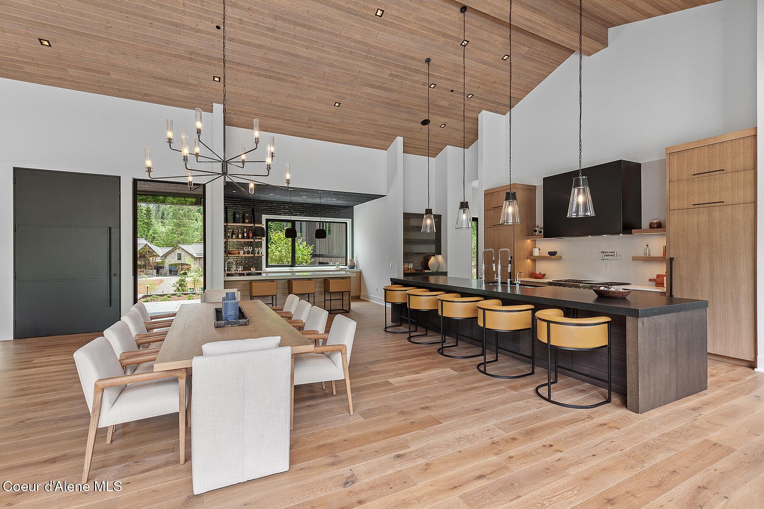 This interior shot showcases a modern, open-concept kitchen and dining area. The kitchen features a large island with bar seating, sleek cabinetry, and pendant lighting, while the dining area includes a long wooden table surrounded by upholstered chairs. The space is characterized by its clean lines, natural wood accents, and abundant natural light, creating a warm and inviting atmosphere.