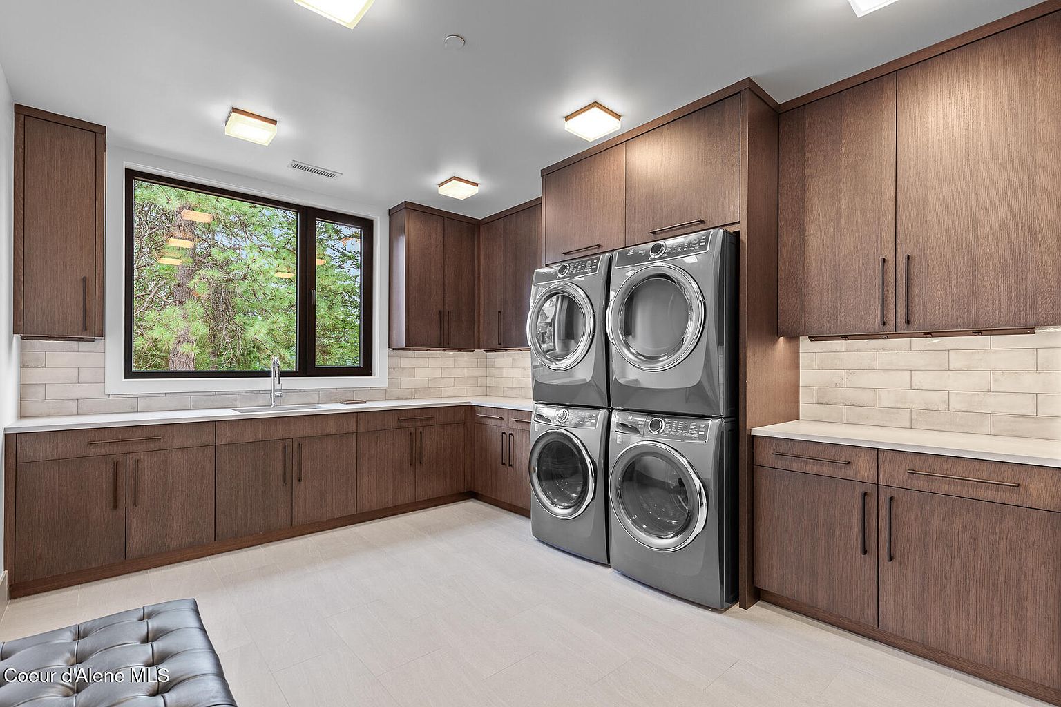 This is a well-appointed laundry room featuring dark wood cabinetry, a white countertop, and stacked washer and dryer units. A large window provides natural light and a view of the outdoors. The room has a clean and modern aesthetic, making laundry chores more enjoyable.
