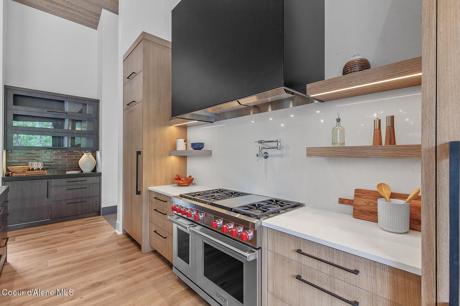 This is a modern kitchen with light wood cabinetry, white countertops, and stainless steel appliances. A black range hood is positioned above the stove, and open shelving with integrated lighting adds a stylish touch. The kitchen has a clean and contemporary design.