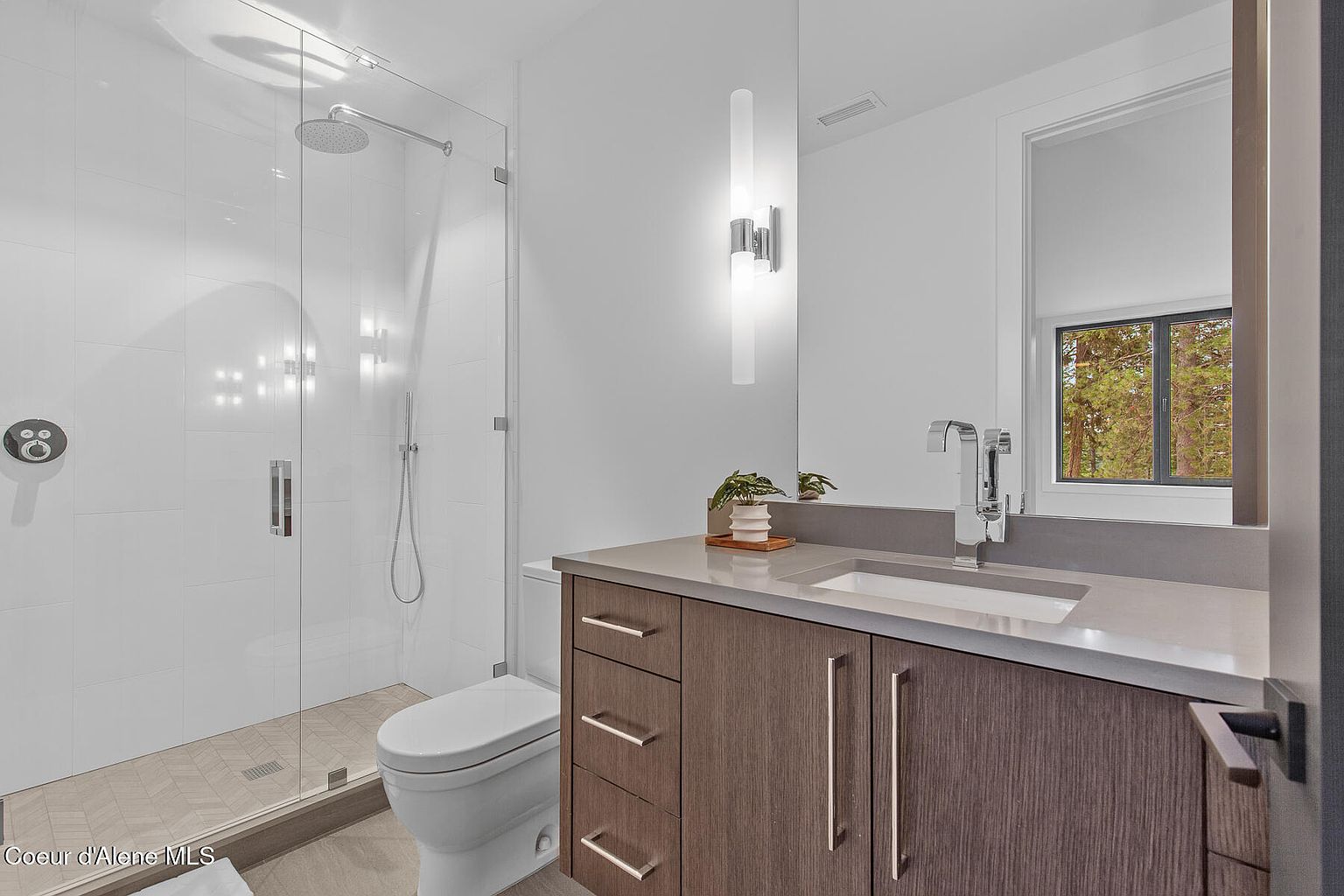This is a modern bathroom featuring a glass-enclosed shower with white tile walls and a rainfall showerhead. A sleek vanity with a gray countertop and dark wood cabinets is complemented by contemporary lighting fixtures. The overall design is clean and minimalist, creating a spa-like atmosphere.
