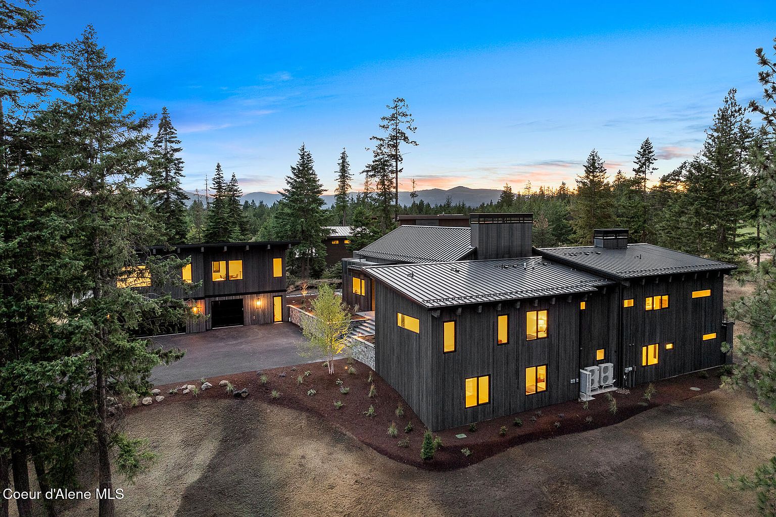 This aerial view showcases a modern, dark-toned home nestled among tall trees, suggesting privacy and a connection with nature. The house features a metal roof and multiple sections, with warm light emanating from the windows, creating an inviting atmosphere. The landscaping is well-maintained, adding to the property's curb appeal.