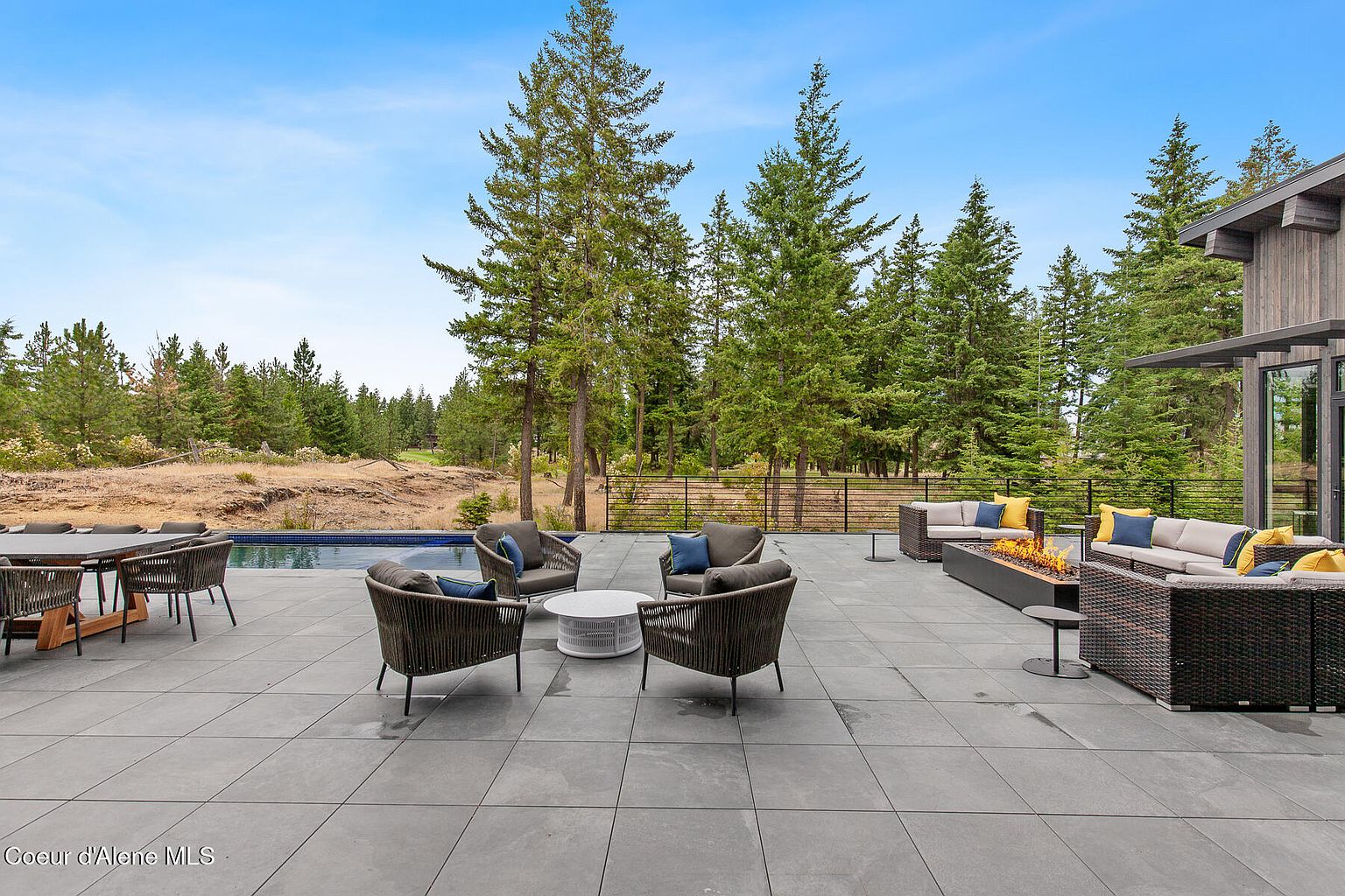 This image showcases a luxurious outdoor patio area, complete with stylish wicker furniture arranged around a modern fire pit. The patio features large format gray tiles, creating a sleek and contemporary look. In the background, there is a swimming pool and lush greenery, enhancing the property's appeal and creating a serene outdoor living space.