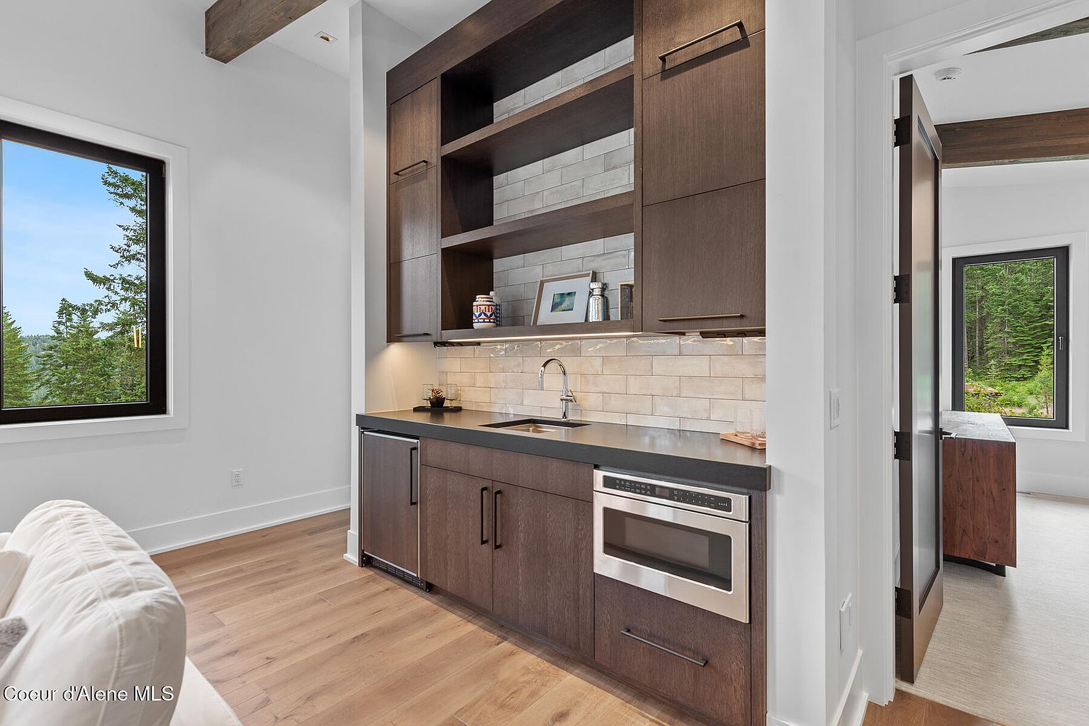 This interior shot showcases a modern living room featuring a wet bar area with dark wood cabinetry, stainless steel appliances, and light-colored subway tile backsplash. A window provides natural light and a view of the outdoors, while the hardwood flooring adds warmth to the space. The room exudes a sophisticated and comfortable ambiance.