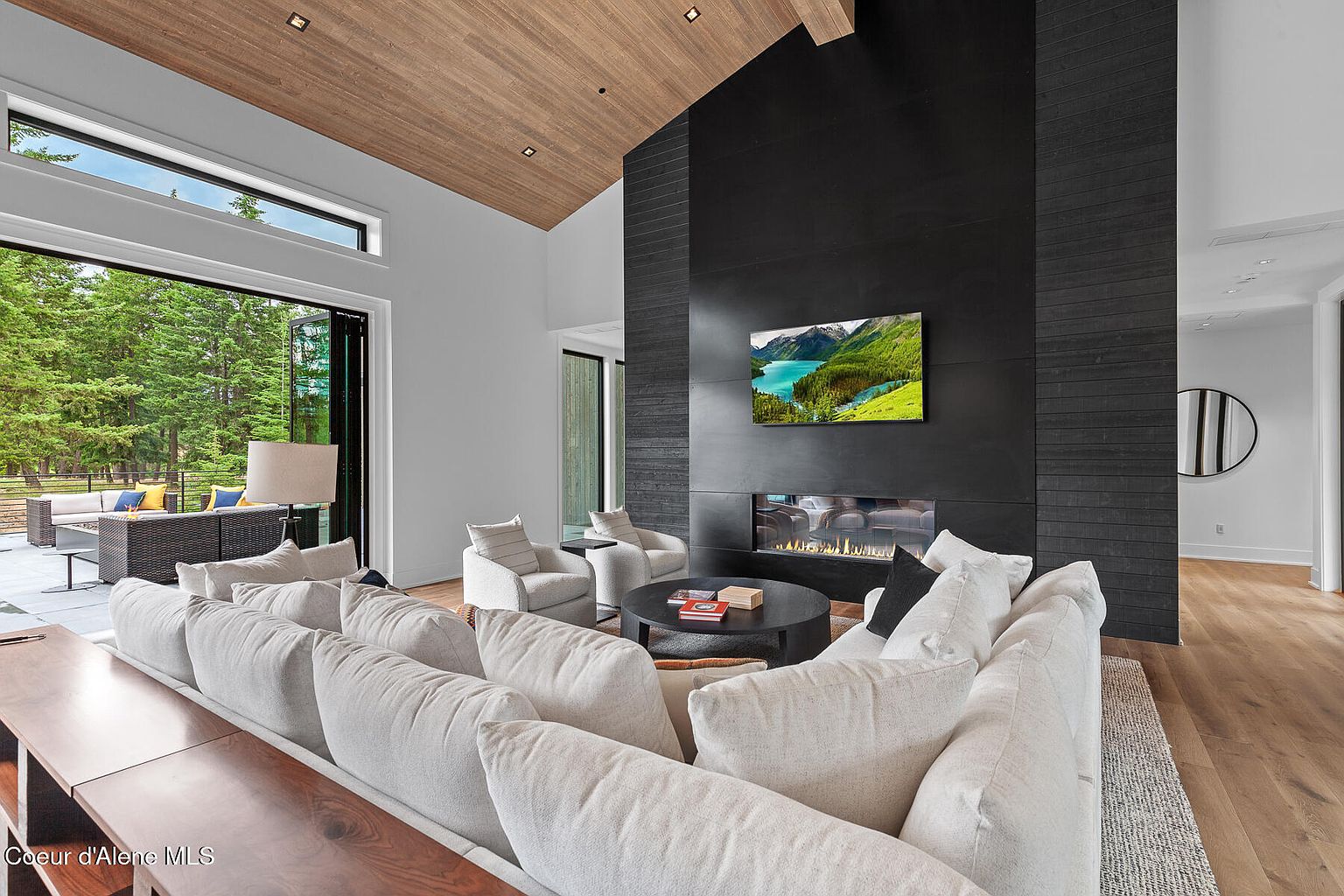This is an interior shot of a modern living room featuring a large, comfortable white sofa and two armchairs arranged around a dark round coffee table. A striking black fireplace with a built-in television dominates one wall, while a large sliding glass door opens to an outdoor patio. The room is well-lit and exudes a sense of contemporary luxury and comfort.