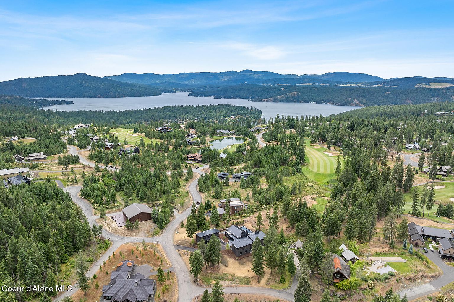 This aerial view showcases a luxurious residential area nestled amidst lush greenery and a serene lake backdrop. The properties feature modern architectural designs with dark roofs, complemented by well-maintained roads and landscaping. A golf course adds to the upscale community's appeal, creating an impression of exclusivity and natural beauty.
