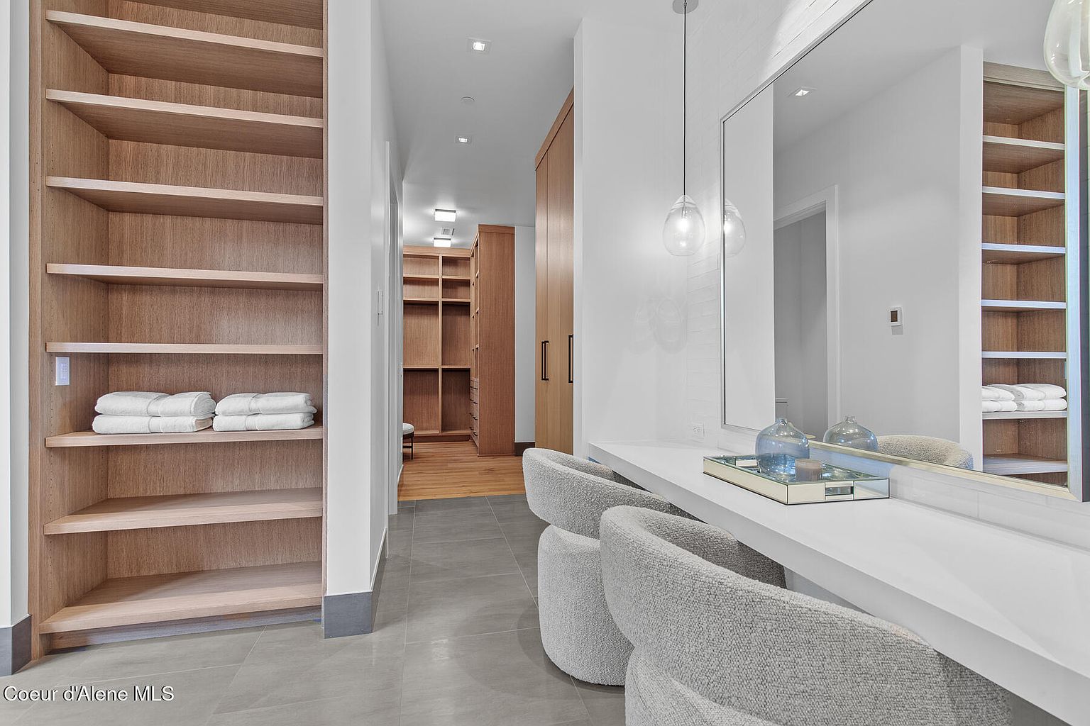 This is an interior shot of a well-organized walk-in closet. The closet features custom wooden shelving units, providing ample storage space for clothing and accessories. A vanity area with a mirror and seating is also present, enhancing the functionality and luxury of the space. The color palette is neutral, creating a clean and sophisticated aesthetic.