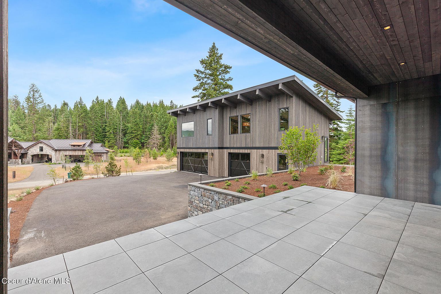 This image showcases the front exterior of a modern home with a striking architectural design. The house features a combination of wood siding and dark metal accents, complemented by a well-manicured landscape. A paved driveway leads to the garage, and a stone retaining wall adds visual interest to the front yard. The overall impression is one of sophisticated elegance and contemporary style.