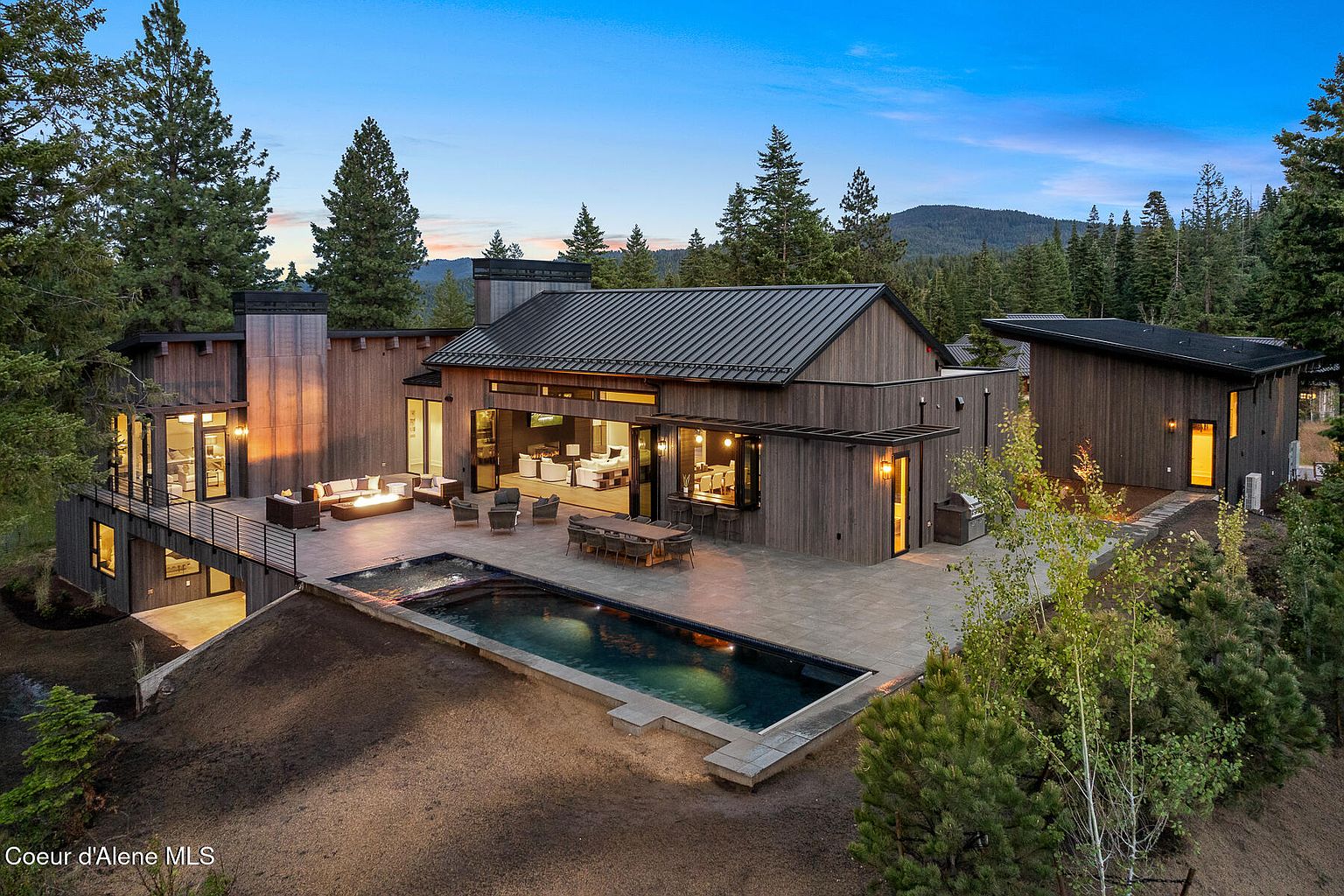 This image showcases the rear exterior of a modern home, emphasizing its outdoor living spaces. A sleek pool is the focal point, complemented by a spacious patio with dining and lounge areas. The architecture features clean lines, dark wood siding, and a metal roof, blending seamlessly with the surrounding trees and mountain backdrop.