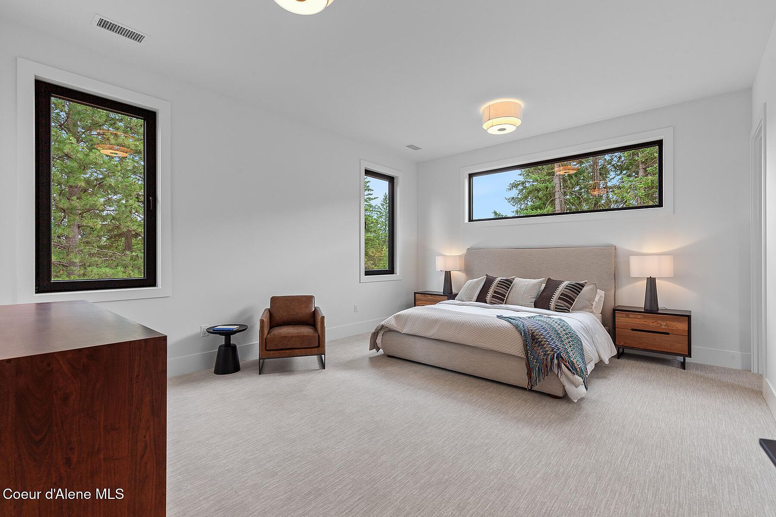 This is a bright and spacious primary bedroom featuring neutral tones and modern design elements. The room includes a large bed with a padded headboard, two nightstands with lamps, and a comfortable armchair. Large windows provide natural light and views of the surrounding trees, enhancing the serene and inviting atmosphere.