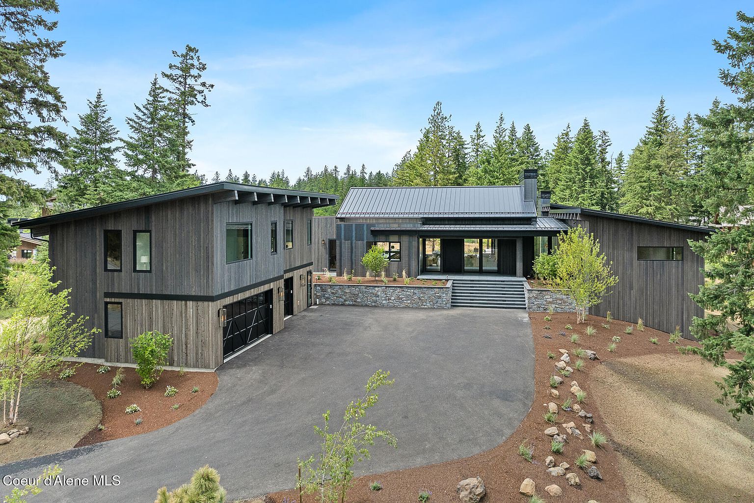 This is a front exterior view of a modern, two-story home with dark wood siding and a metal roof. The house features a two-car garage on the left and a landscaped front yard with stone accents and young trees. A wide driveway leads up to the house, and the surrounding area is wooded, providing privacy.