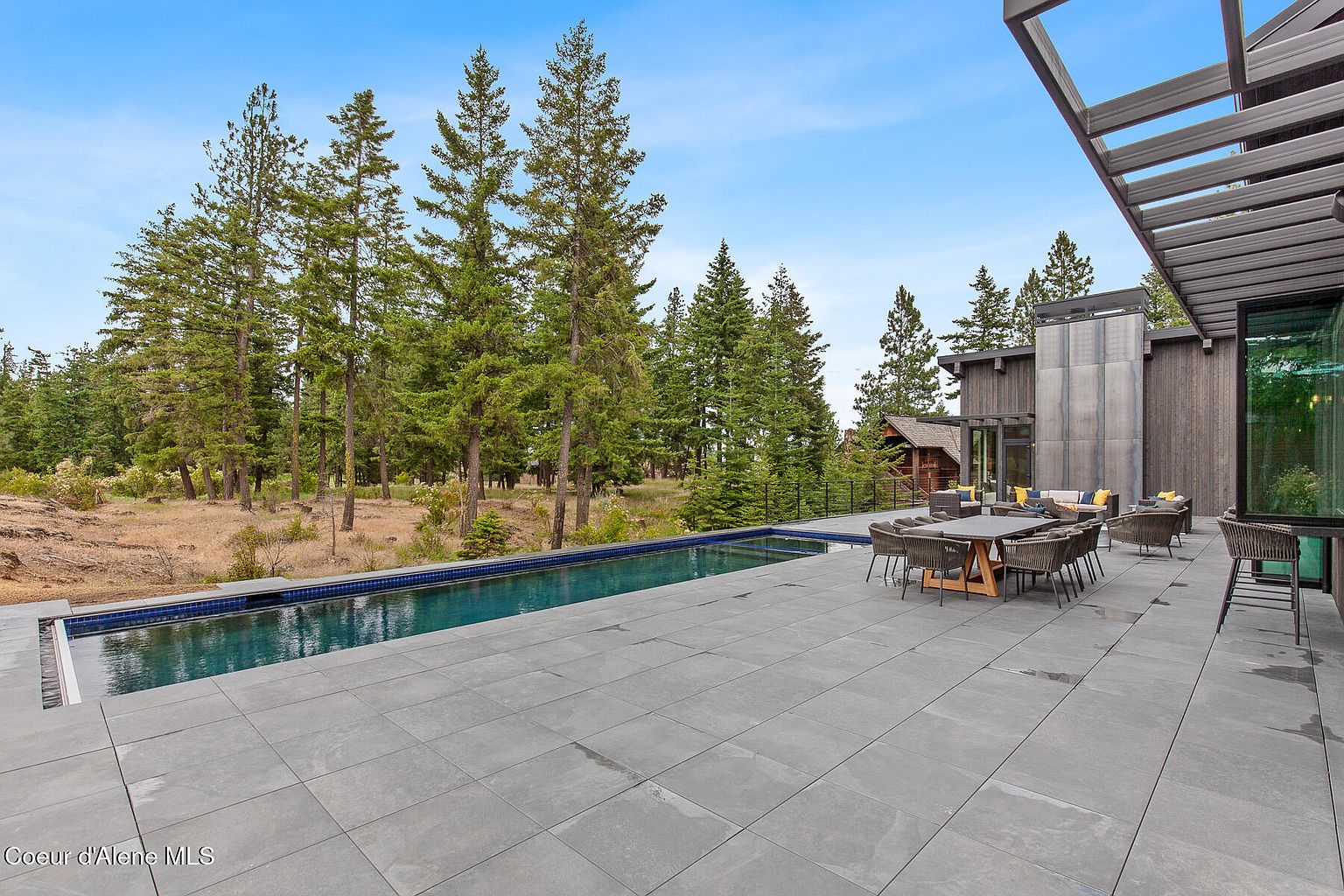 This image showcases a luxurious outdoor space featuring a sleek, modern pool with dark blue water, surrounded by a spacious gray tiled patio. An outdoor dining set and lounge area are positioned near the house, which boasts a contemporary design with wood and metal accents. Tall pine trees provide a scenic backdrop, enhancing the property's appeal.