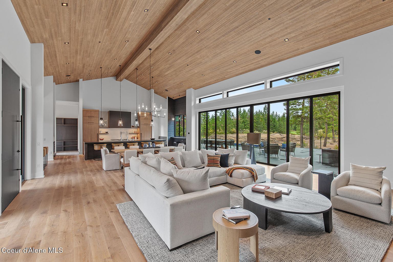 This is a spacious and modern living room featuring a large sectional sofa, two armchairs, and a round coffee table on a neutral-toned rug. The room has a high, wood-paneled ceiling and large sliding glass doors that offer a view of the outdoor patio and surrounding trees. The open-concept design seamlessly connects the living area to the kitchen, creating a cohesive and inviting space.