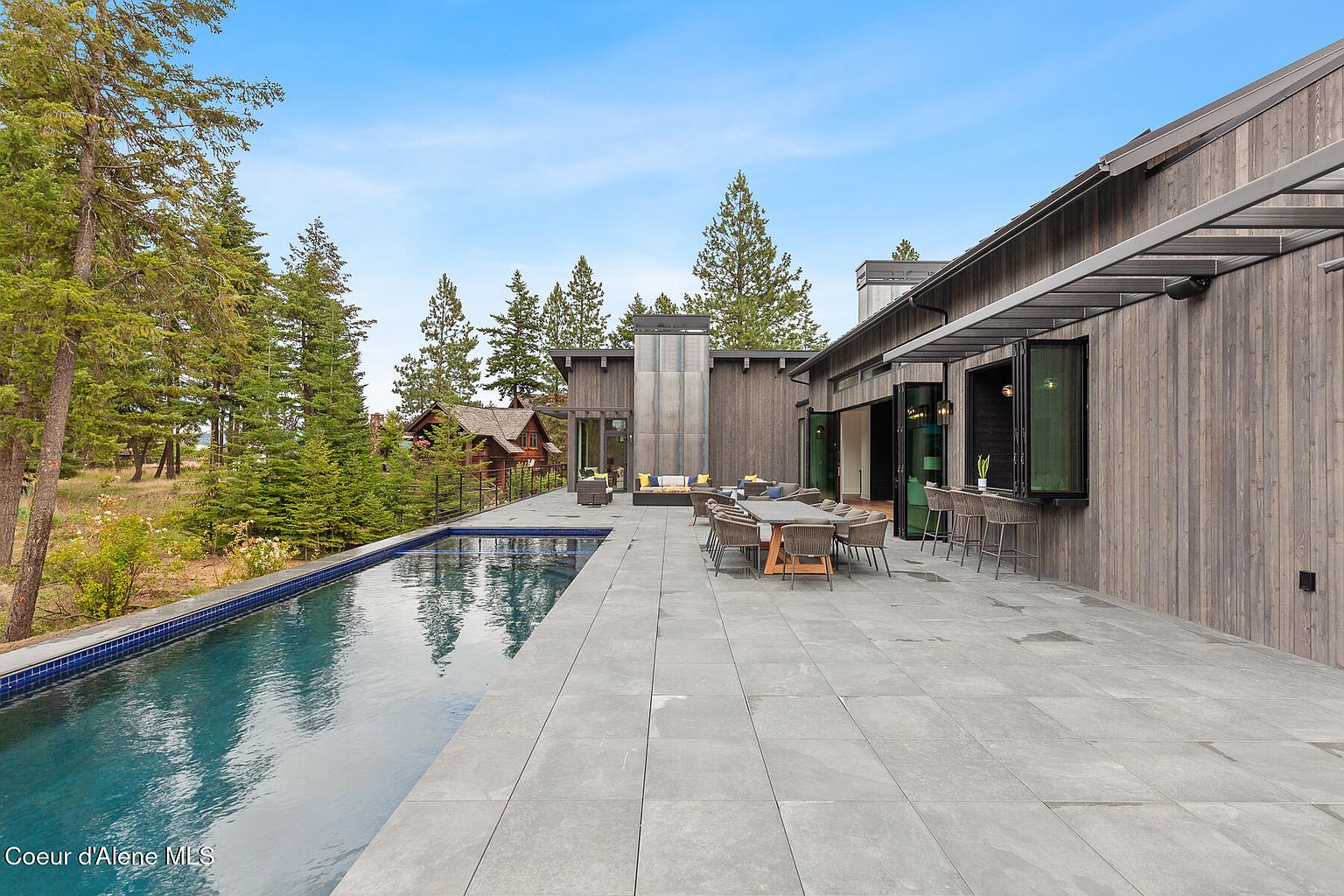 This image showcases a luxurious outdoor space featuring a modern home with a long, rectangular pool. The pool is surrounded by a spacious patio with outdoor dining and lounge areas, perfect for entertaining. The architecture is contemporary with clean lines and natural wood siding, complemented by lush greenery and mature trees in the background, creating a serene and private setting.