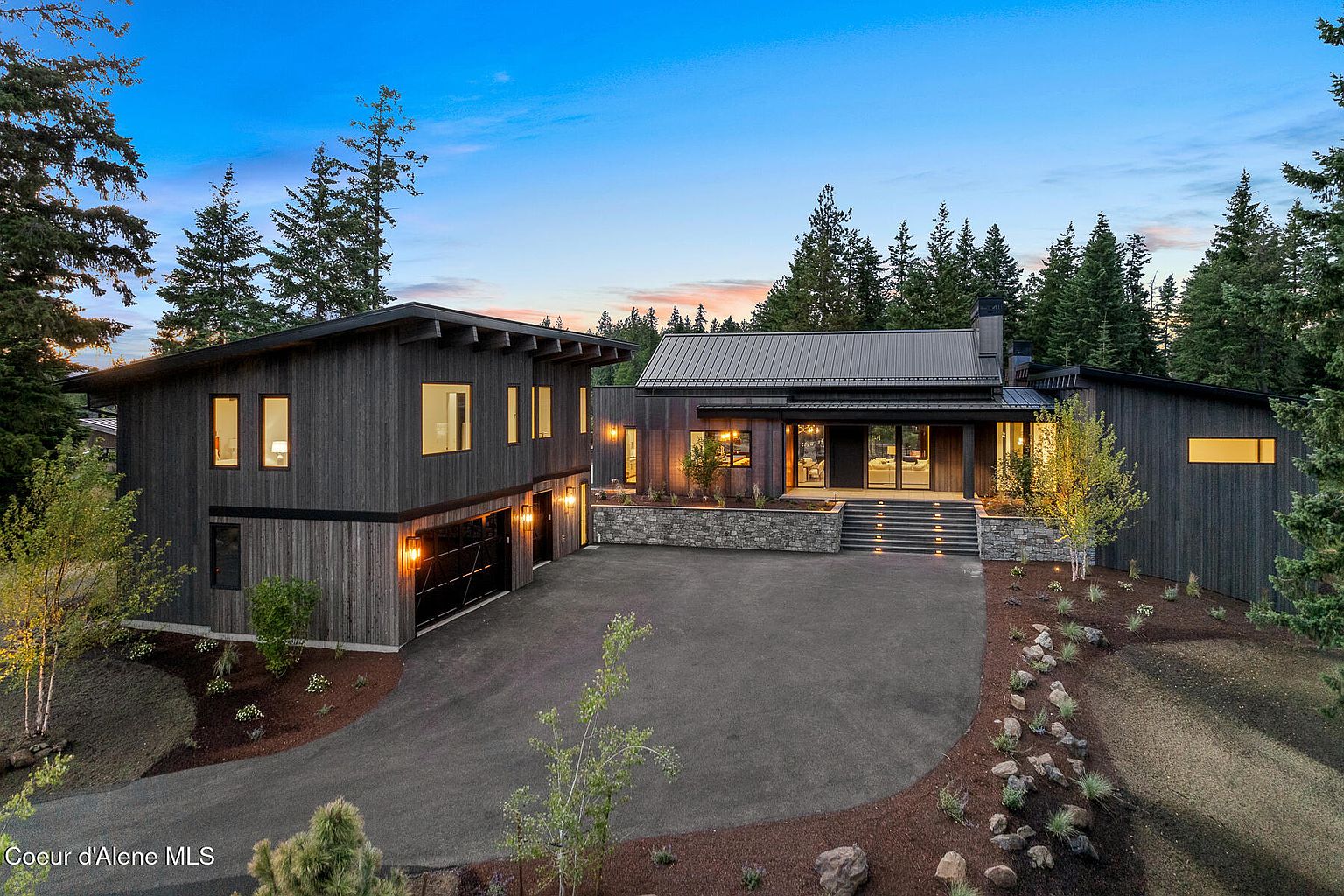 This is a front exterior view of a modern home with dark wood siding and a metal roof. The house features a two-car garage on the left and a main entrance with stone steps leading up to the front door. The driveway is paved and surrounded by landscaping with trees and shrubs, creating a serene and private setting.