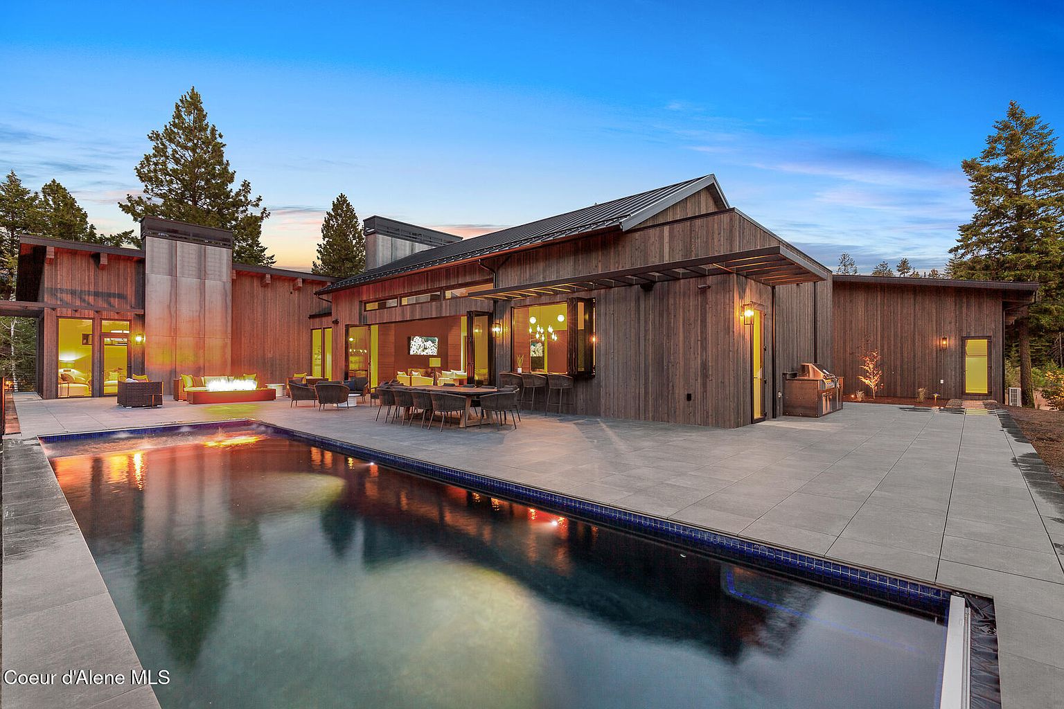 This image showcases the rear exterior of a modern home, emphasizing the luxurious pool and patio area. The architecture features clean lines, wood siding, and a dark metal roof. Outdoor seating and a fire pit create an inviting space for relaxation and entertainment, while the pool's reflective surface adds to the serene ambiance.