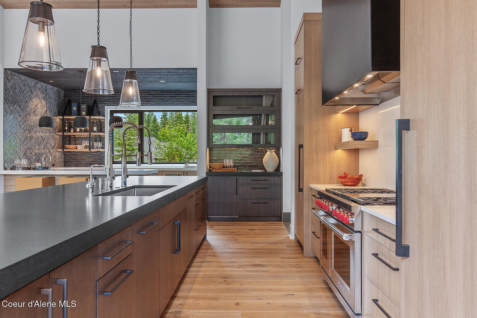 This is a modern kitchen featuring dark wood cabinets and a large island with a dark countertop. Stainless steel appliances are visible, including a range with red knobs and a black range hood. The flooring is light wood, and pendant lights hang over the island, creating a warm and inviting atmosphere.