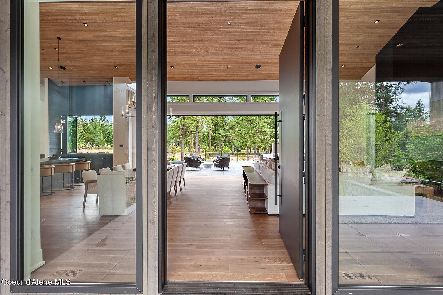 This interior shot showcases a modern living room viewed through a grand entryway. The open floor plan leads to an outdoor patio with seating, framed by lush greenery. The interior features hardwood floors, a sleek console table, and contemporary furnishings, creating a seamless transition between indoor and outdoor living spaces.
