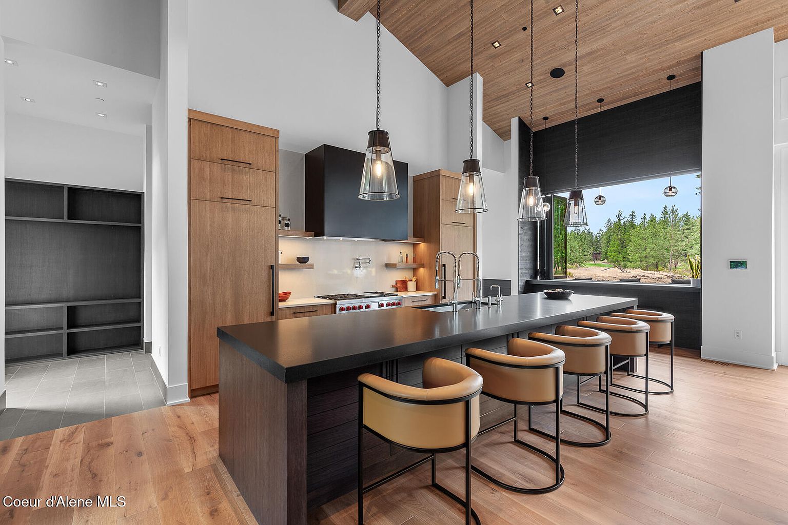 This is a modern kitchen with wood cabinets, a black range hood, and a dark countertop island with seating. The kitchen features pendant lighting and a large window offering a view of the outdoors. The overall style is contemporary and luxurious.