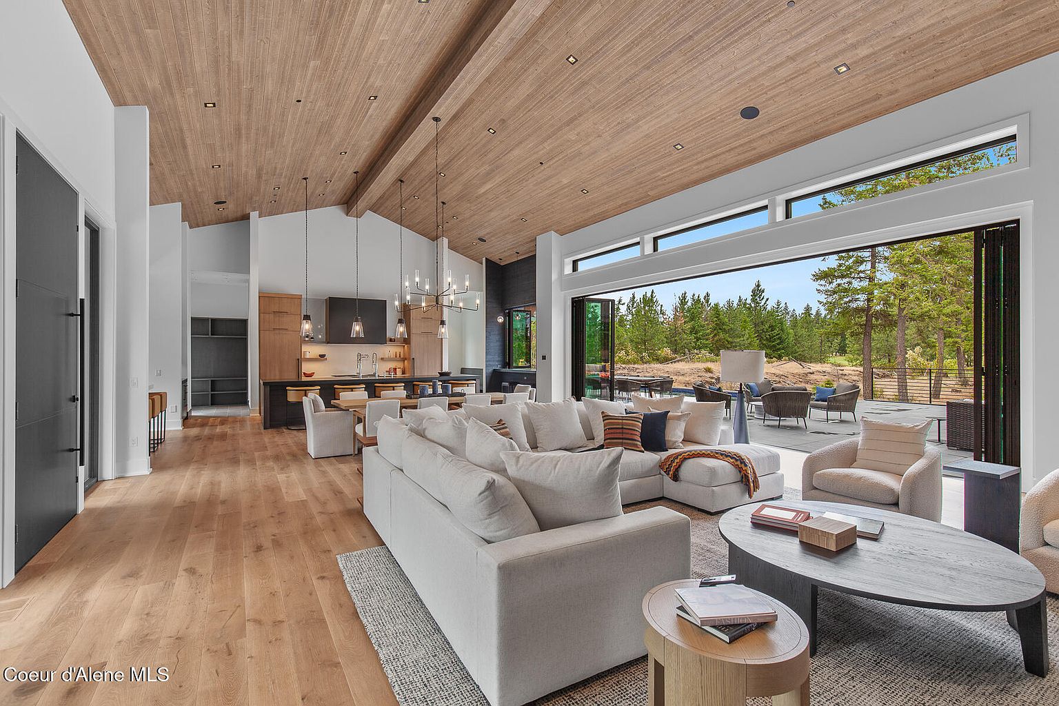 This is an interior shot of a modern living room that seamlessly blends indoor and outdoor living. The room features a large sectional sofa, a wooden ceiling with recessed lighting, and an open floor plan that connects to the kitchen. A large sliding glass door opens to an outdoor patio area, providing ample natural light and a view of the surrounding landscape.