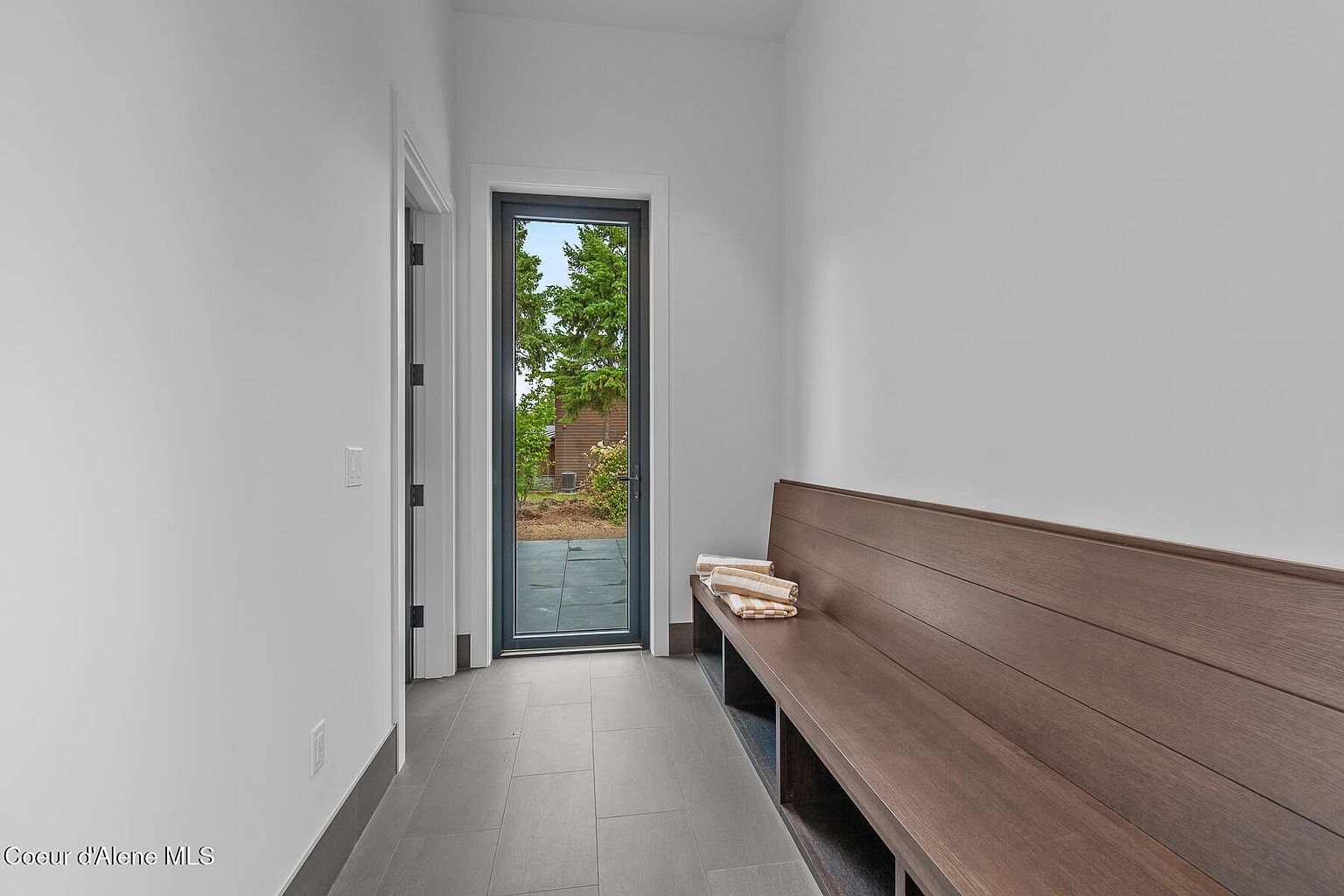 This interior shot showcases a modern hallway with gray tile flooring and white walls. A long wooden bench sits against the right wall, topped with neatly folded towels. A glass door at the end of the hallway provides a view of the exterior greenery, enhancing the sense of space and light.