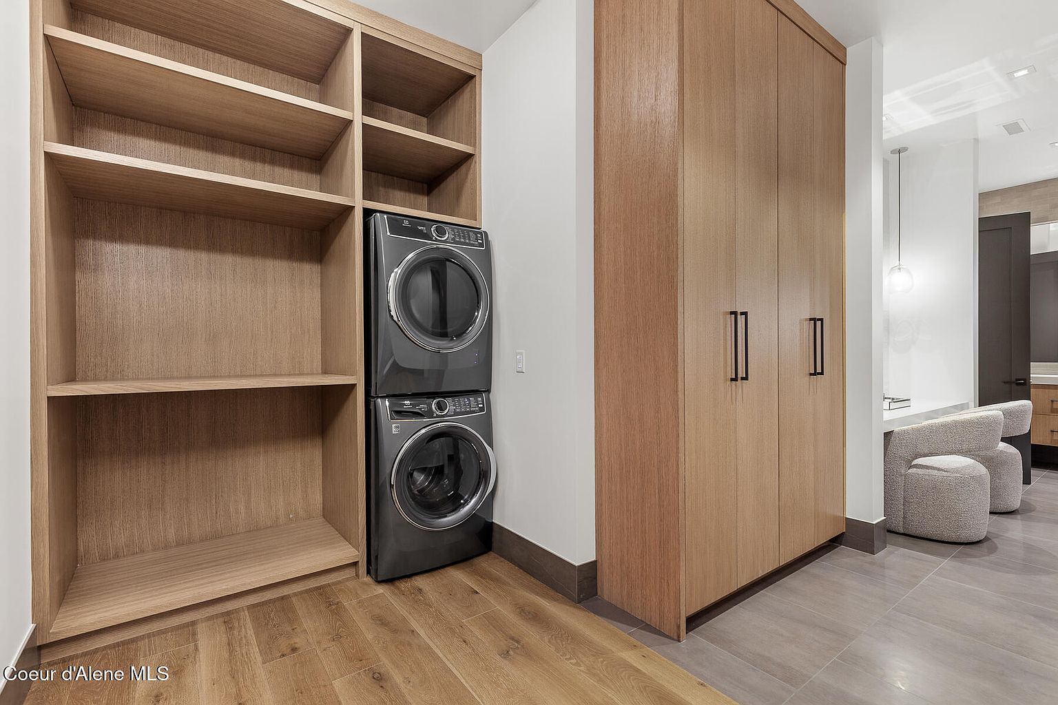 This is an interior shot of a laundry room featuring a stacked washer and dryer unit nestled between built-in wooden shelves. To the right, a tall wooden cabinet with sleek, dark handles adds storage. The flooring transitions from wood to tile, leading to a glimpse of a seating area, suggesting a well-designed and functional space.