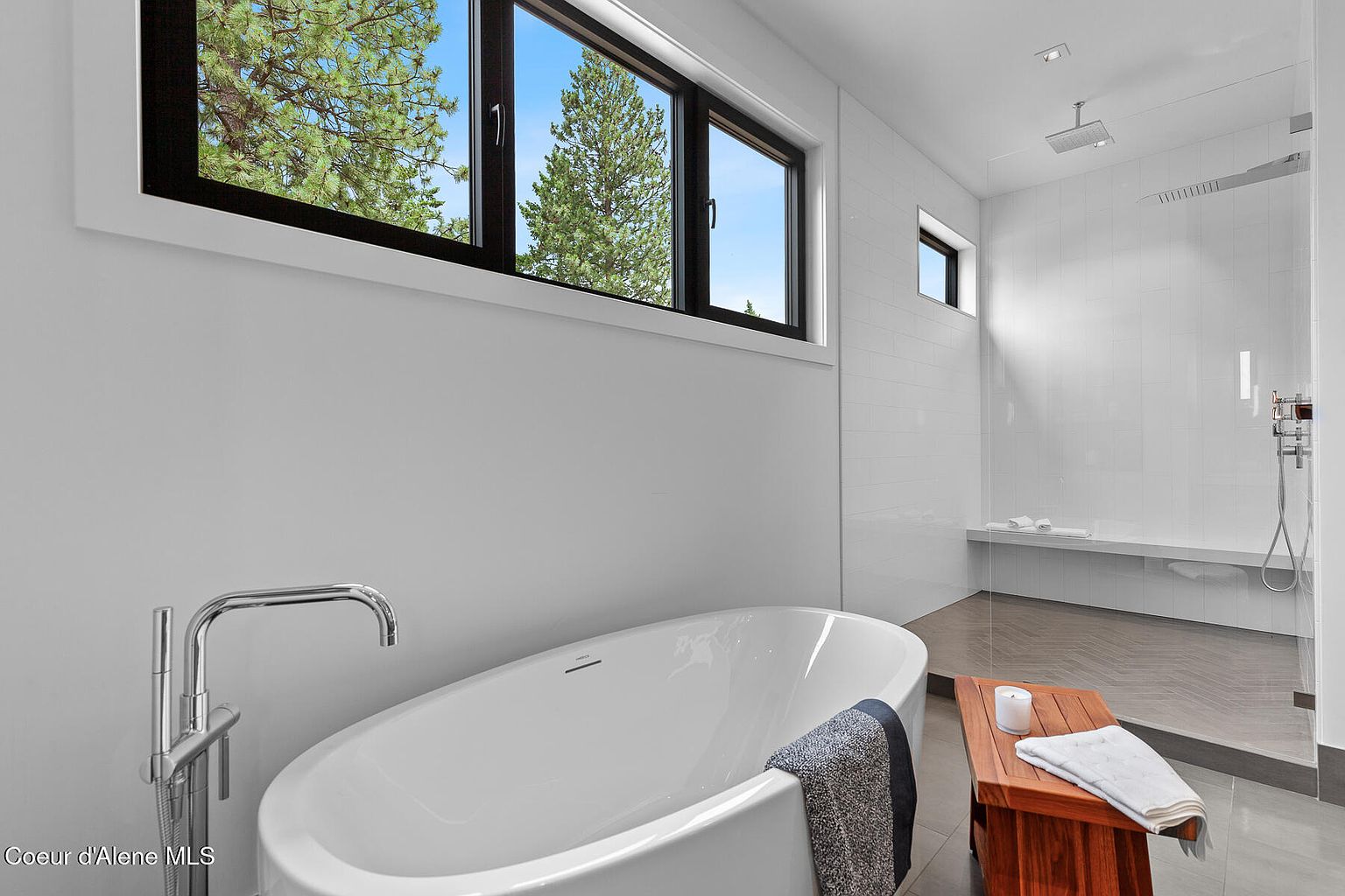 This is a bright and modern primary bathroom featuring a freestanding white bathtub with a chrome faucet. A large window offers a view of trees, and a walk-in shower with a bench is visible in the background. The bathroom has white walls and tile, with a wooden bench adding a touch of warmth.