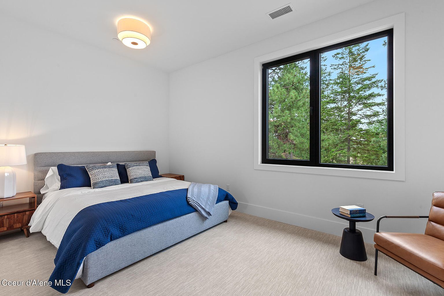 This is a bright and airy bedroom featuring a gray upholstered bed with blue and patterned pillows, a white duvet, and a blue throw blanket. A modern black-framed window offers a view of tall green trees, and a small black side table with books sits next to a leather chair. The room has a minimalist aesthetic with white walls and light-colored carpet, creating a serene and inviting atmosphere.