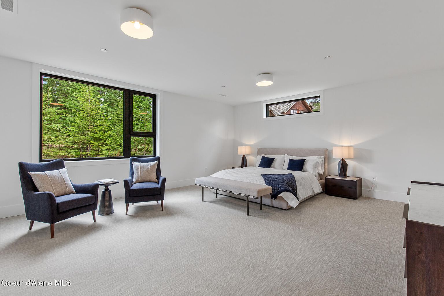 This is a spacious primary bedroom featuring a large window with a view of lush greenery. The room is decorated in a minimalist style with white walls, light gray carpet, and contemporary furniture. A king-size bed with neutral bedding and a bench at the foot adds to the serene and inviting atmosphere.