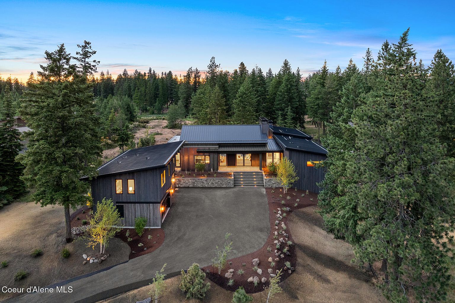 This aerial view showcases a modern, dark-colored home nestled among lush trees. The property features a well-maintained driveway, professional landscaping, and a striking architectural design with a dark roof. The surrounding forest provides a sense of privacy and seclusion, enhancing the property's appeal.