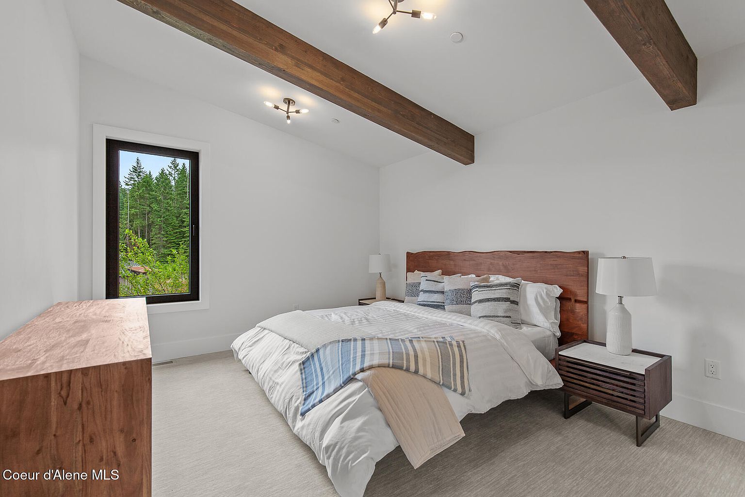 This is a bright and airy bedroom featuring a bed with a wooden headboard, white linens, and decorative pillows. The room has white walls, wooden beams on the ceiling, and a large window offering natural light and a view of the outdoors. A wooden nightstand with a lamp sits beside the bed, and a wooden dresser is visible on the left.