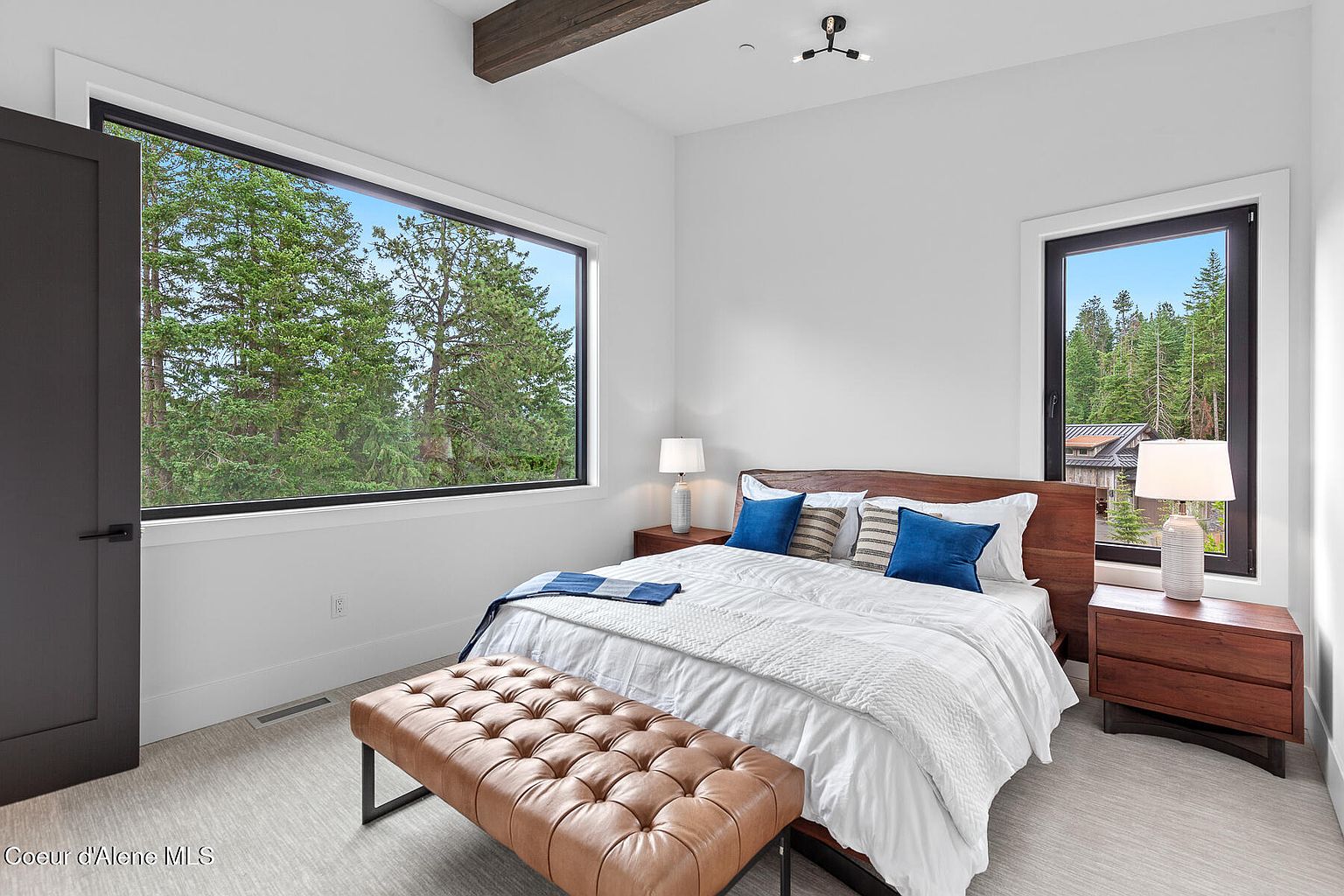 This is a bright and airy primary bedroom featuring a wooden bed frame with white linens and blue accent pillows. Large windows offer views of the surrounding trees, enhancing the room's natural light. The room is furnished with wooden nightstands and a tufted leather bench, creating a cozy and inviting atmosphere.
