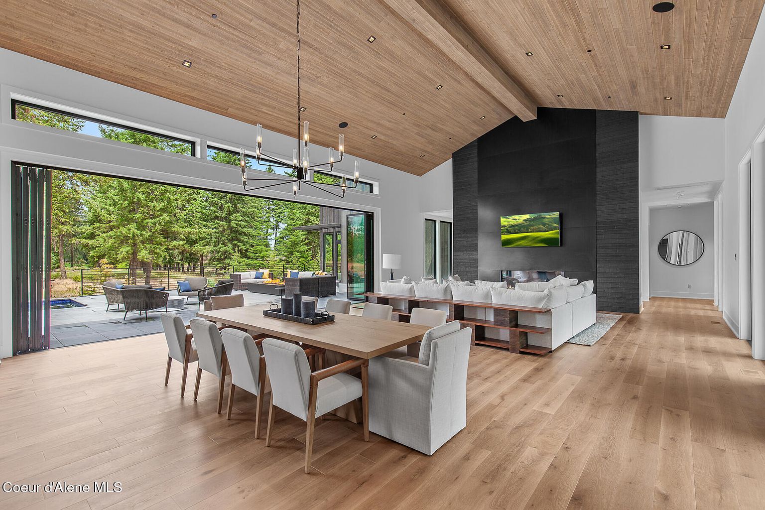 This is a spacious and modern living room with an open floor plan, seamlessly blending indoor and outdoor living. The room features a dining area with a large wooden table and white chairs, a comfortable seating area with a white sectional sofa, and a striking black fireplace wall with a mounted television. Large sliding glass doors open to an outdoor patio area, offering views of the surrounding landscape.