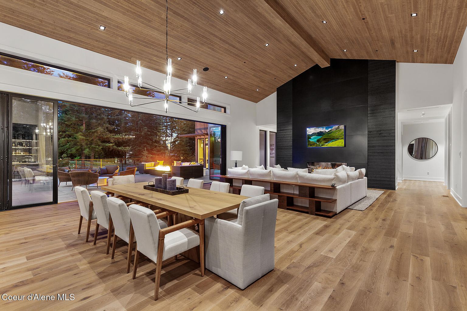 This is an interior shot of a luxurious living and dining area. The space features a modern chandelier, a long wooden dining table with upholstered chairs, and a spacious living area with a large sectional sofa facing a black accent wall with a television. Large sliding glass doors open to an outdoor patio with a fire pit, creating a seamless indoor-outdoor living experience.