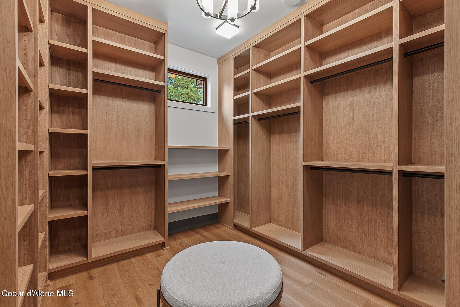 This is a well-organized walk-in closet featuring custom wooden shelving and hanging rods. The closet is spacious and well-lit, with a neutral color palette and a round ottoman in the center. The hardwood flooring adds a touch of elegance to the space.