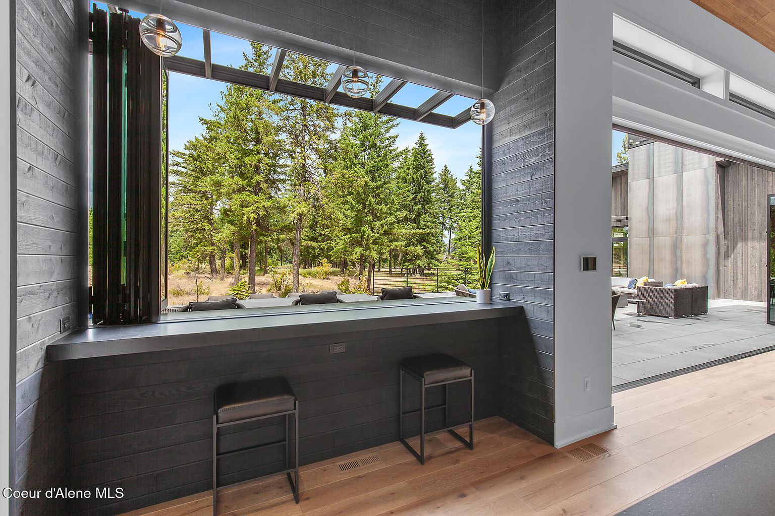This interior shot showcases a modern living space with a bar area overlooking a lush green landscape through expansive windows. The bar features dark wood paneling and sleek stools, while the open layout seamlessly connects to an outdoor patio area. The hardwood flooring and contemporary lighting fixtures add to the sophisticated ambiance of the room.