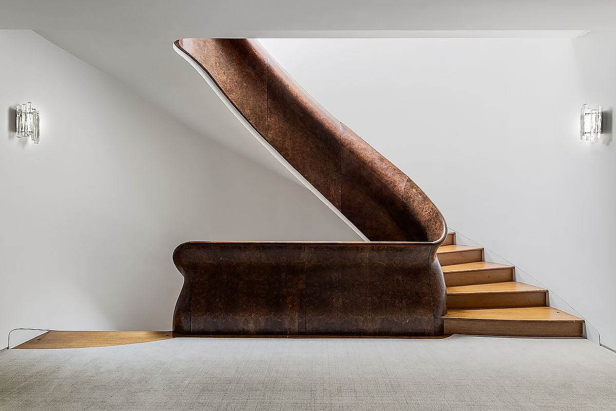 This image captures a striking staircase featuring a curved, dark-toned handrail and balustrade that creates a sculptural focal point against the minimalist white walls. The wooden steps lead upward, complemented by elegant wall-mounted light fixtures on either side. The perspective is centered and symmetrical, emphasizing the clean lines and sophisticated architectural design of the space.