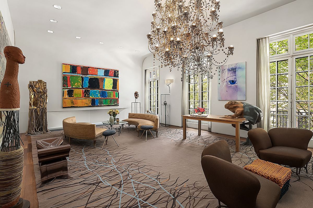 This expansive and sophisticated living room features a dramatic crystal chandelier as its centerpiece, hanging above a large, patterned area rug. The space is decorated with eclectic art pieces, including a large abstract painting and a sculptural frog figure on a wooden console table. Tall windows with sheer curtains allow natural light to filter in, creating an airy and gallery-like atmosphere.