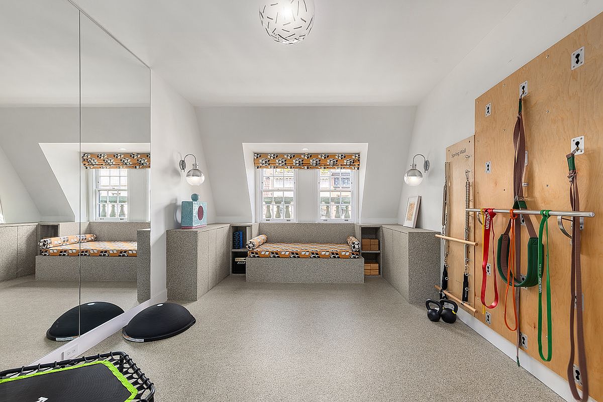 This dedicated home gym features a wall-mounted spring exercise system, a large mirrored wall, and a built-in bench seat with patterned upholstery beneath a dormer window. The room is finished with neutral carpeting and soft, recessed lighting, creating a functional and organized space for fitness. The perspective is a wide-angle shot from the center of the room, capturing the full layout of the workout equipment and seating area.