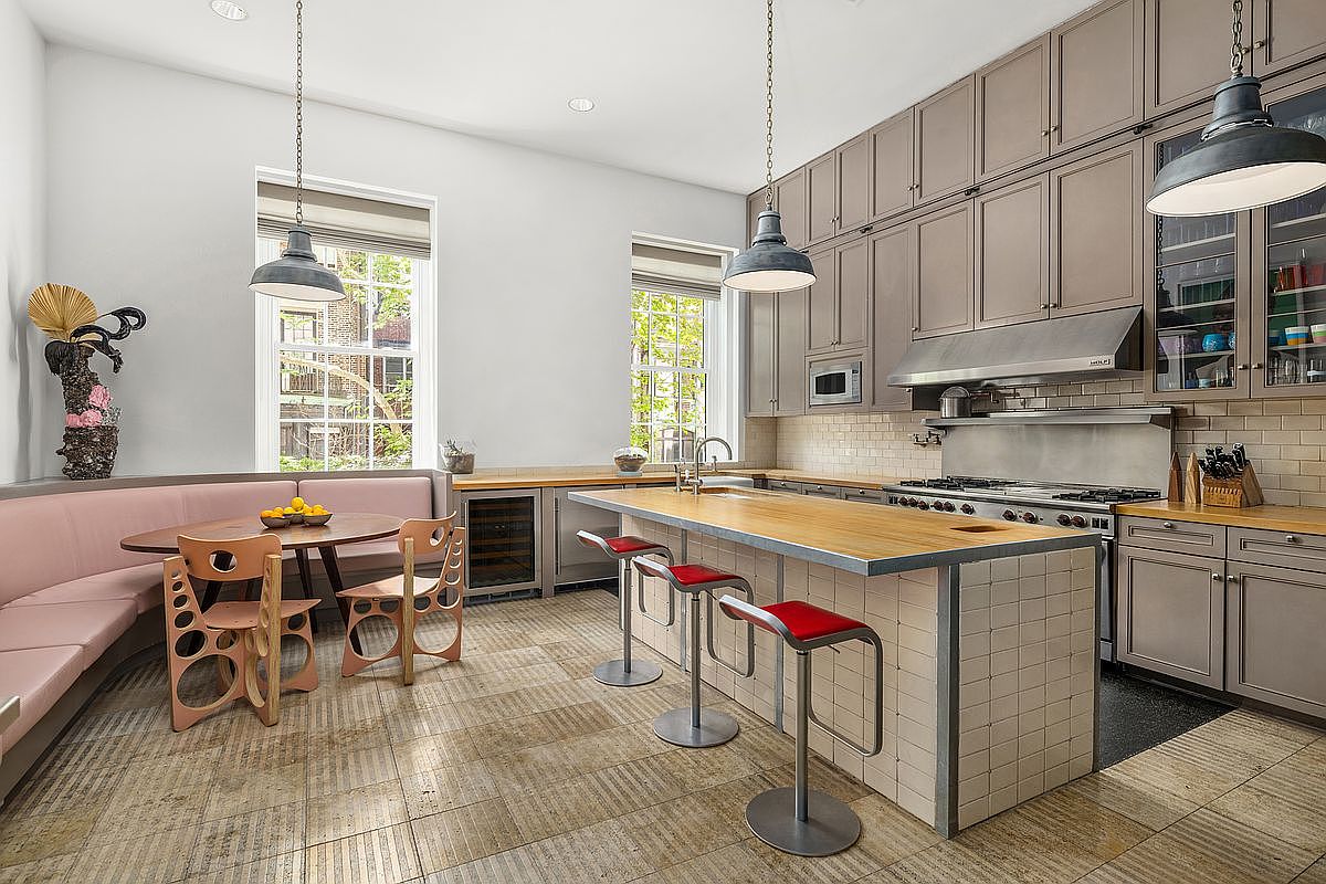 This spacious, modern kitchen features a large central island with a light wood countertop and red-cushioned bar stools, complemented by a matching dining nook with a built-in pink banquette. The cabinetry is finished in a muted taupe, paired with stainless steel appliances and a professional-grade range hood. Large windows allow natural light to flood the room, highlighting the textured tile flooring and industrial-style pendant lighting.