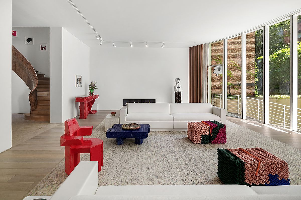 This spacious, modern living room features a minimalist aesthetic with white walls, light wood flooring, and a large area rug. The room is anchored by a white sofa and accented with bold, sculptural furniture pieces in vibrant red and blue, alongside textured, woven ottomans. A wall of floor-to-ceiling windows provides abundant natural light and a view of the exterior brick wall and greenery, while a curved wooden staircase adds an architectural focal point to the left.