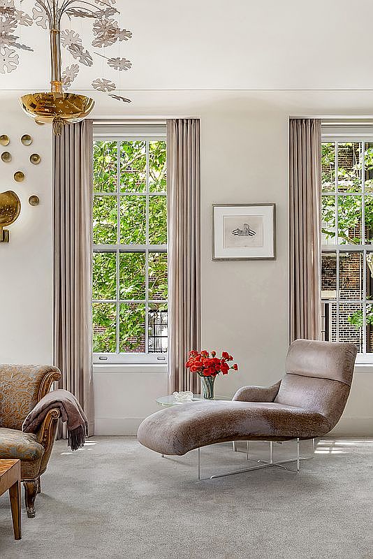 This elegant living room features a sophisticated, neutral color palette with a plush chaise lounge and a patterned armchair. Tall windows draped in soft, floor-length curtains allow natural light to filter in, highlighting the textured carpet and a unique, sculptural chandelier. The space is accented by a small glass-topped table with a vibrant bouquet of red flowers and a framed piece of art on the wall, creating a refined and tranquil atmosphere.