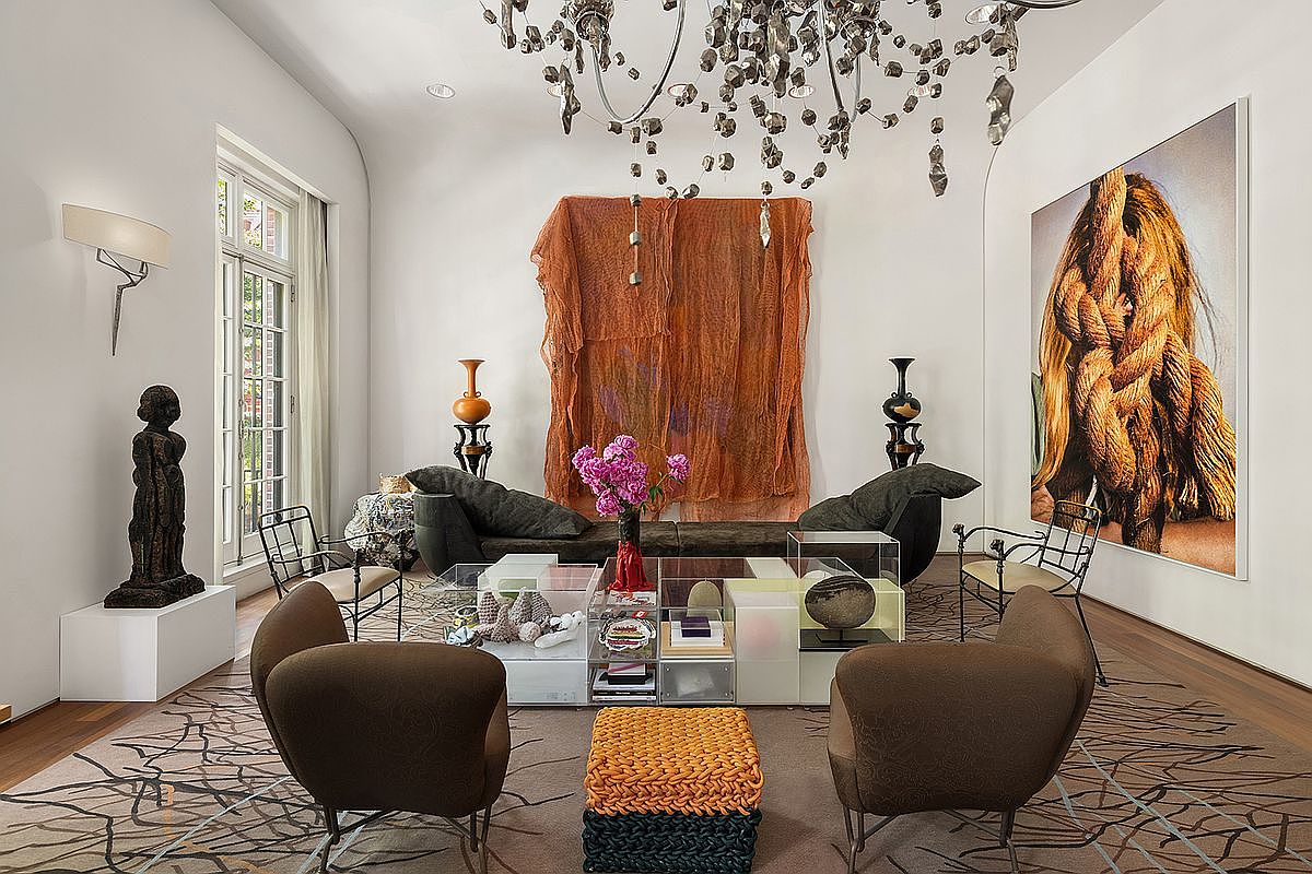 This sophisticated living room features a high-ceiling design with a dramatic, multi-tiered metallic chandelier as a central focal point. The space is furnished with a low-profile sofa, two rounded armchairs, and a unique modular coffee table, all set upon a patterned area rug. A large, textured wall hanging and a striking piece of wall art add artistic flair, while tall windows with sheer curtains allow natural light to filter into the room.