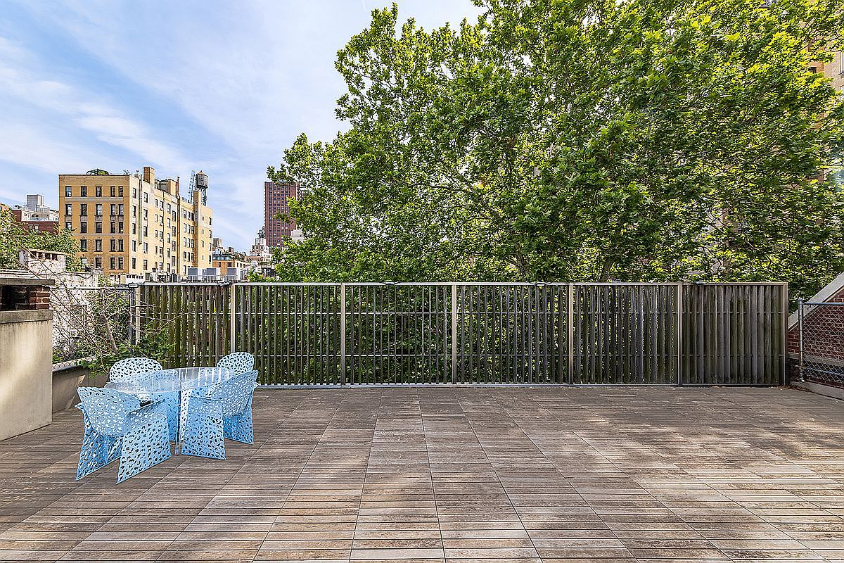 This spacious rooftop deck features light-toned wood decking and a modern, slatted metal privacy screen that overlooks a lush, mature tree. A set of distinctive, perforated blue chairs surrounds a glass-topped table, creating a vibrant seating area. The perspective is eye-level, capturing the urban setting with surrounding city buildings visible in the background under a clear sky.