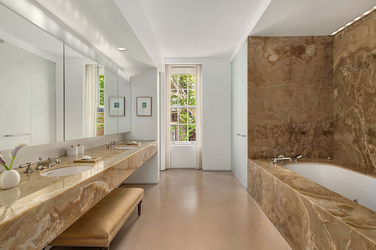 This luxurious primary bathroom features extensive use of warm-toned, veined stone on the vanity countertop and the bathtub surround. A long, wall-to-wall mirror reflects the space, enhancing the sense of depth, while a bench seat sits neatly beneath the vanity. The room is anchored by a deep soaking tub and illuminated by soft, recessed lighting, creating a serene and sophisticated atmosphere.