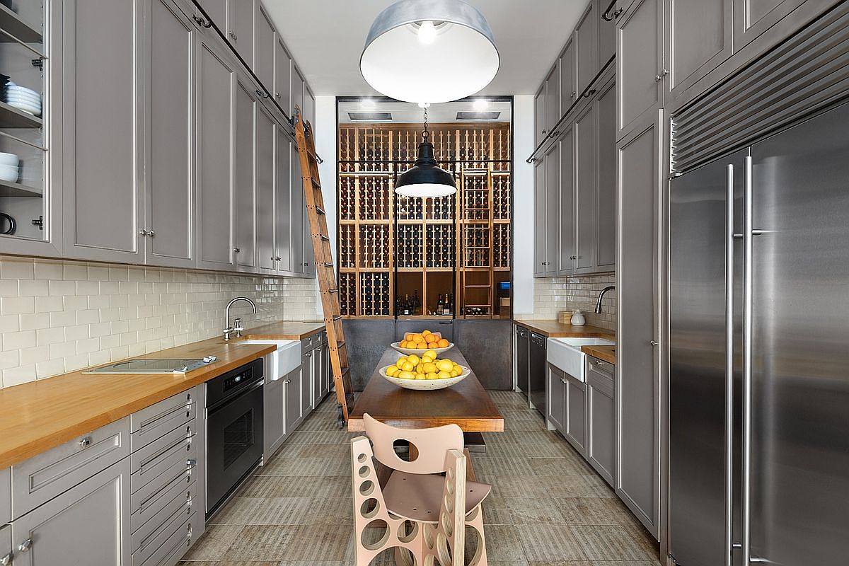 This sophisticated galley-style kitchen features extensive grey cabinetry, warm wood countertops, and a central dining table topped with bowls of fresh fruit. A rolling library-style ladder provides access to the high upper cabinets, while a large, integrated wine storage wall serves as a dramatic focal point at the end of the room. The space is illuminated by a large industrial-style pendant light, creating a refined and functional culinary environment.