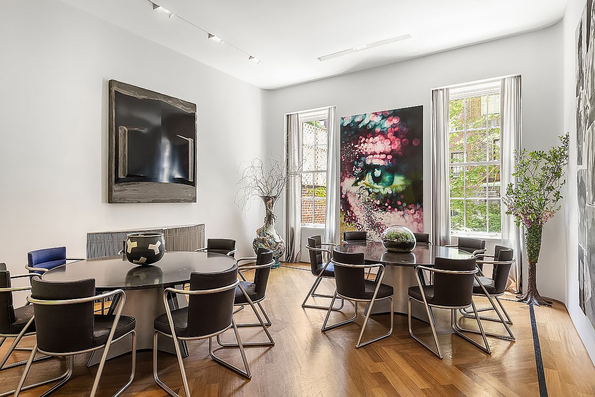 This sophisticated dining room features two large, dark-toned oval tables, each surrounded by modern black leather chairs with sleek metal frames. The space is adorned with bold, contemporary artwork, including a striking close-up portrait of an eye and a large abstract piece, while tall windows draped in sheer curtains allow natural light to fill the room. The polished wood flooring and minimalist aesthetic create an elegant, gallery-like atmosphere suitable for formal entertaining.