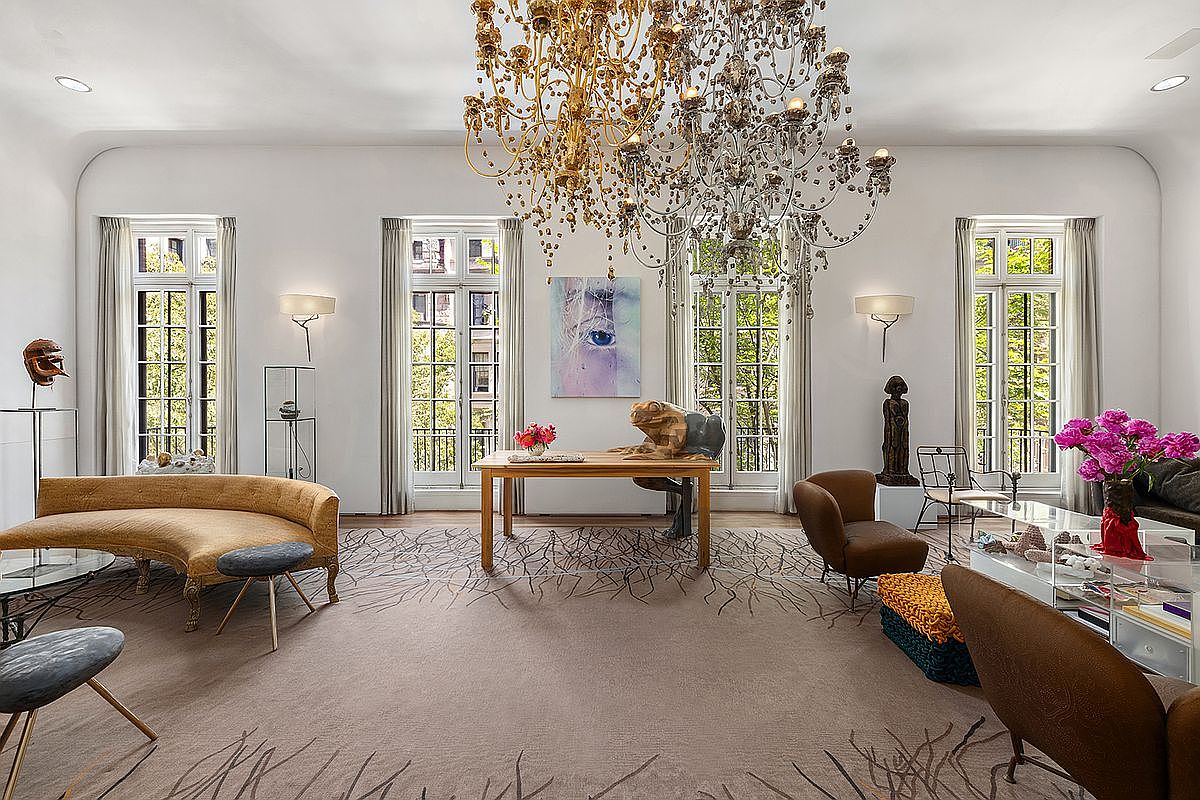 This sophisticated living room features a grand, ornate crystal chandelier as its centerpiece, hanging above a central wooden table adorned with a unique frog sculpture. The space is furnished with a curved, mustard-toned sofa and plush armchairs, all set upon a large, neutral-toned rug with a delicate, dark-lined branch pattern. Tall, multi-paned windows line the walls, allowing ample natural light to fill the room, while curated art pieces and decorative accents contribute to a refined, gallery-like atmosphere.