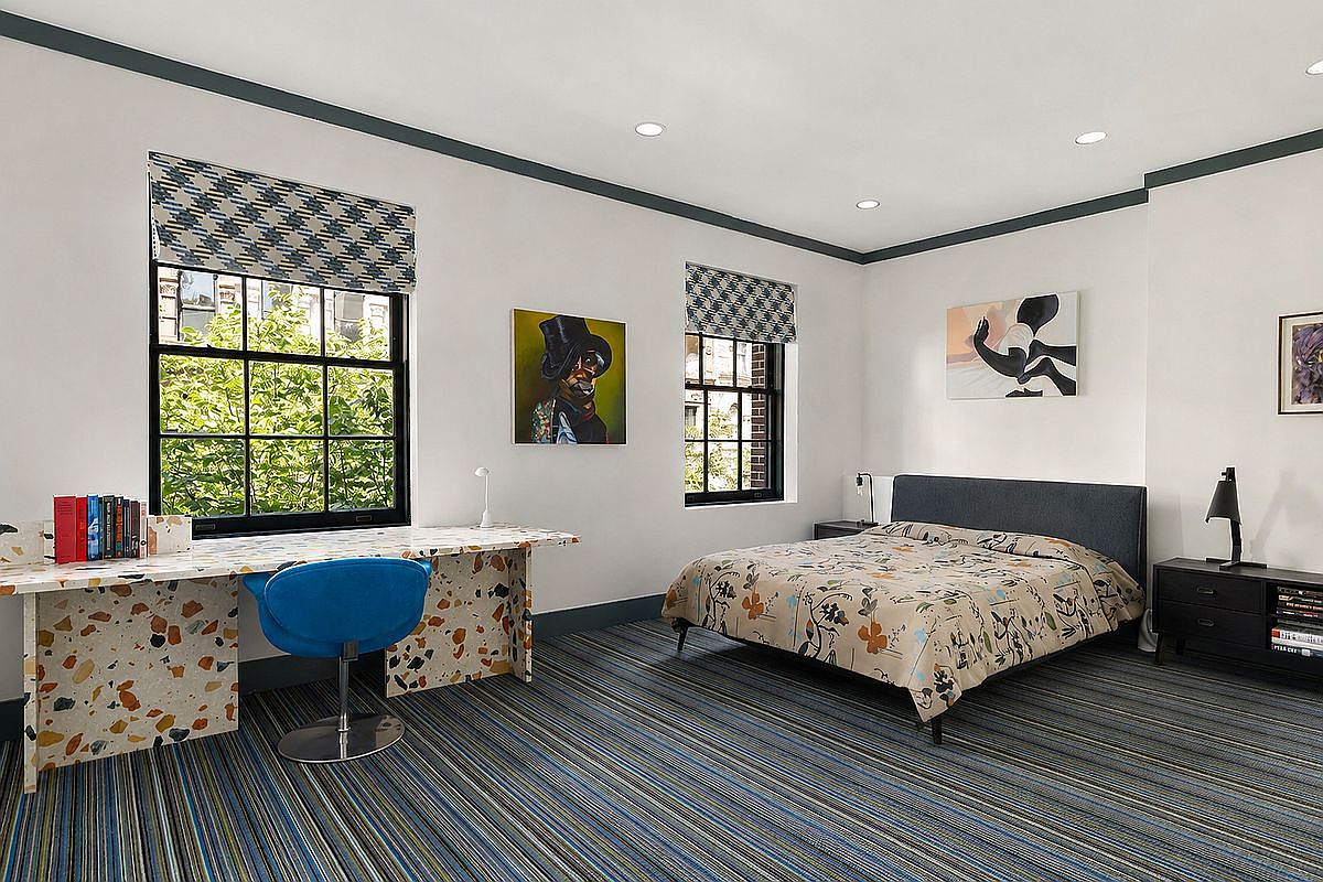 This contemporary bedroom features a unique terrazzo-style desk integrated into the wall, paired with a vibrant blue swivel chair. The room is anchored by a bed with a patterned duvet and a dark upholstered headboard, complemented by a sleek black nightstand and modern artwork. Large windows with patterned shades allow natural light to fill the space, which is finished with bold, striped carpeting and dark trim accents.