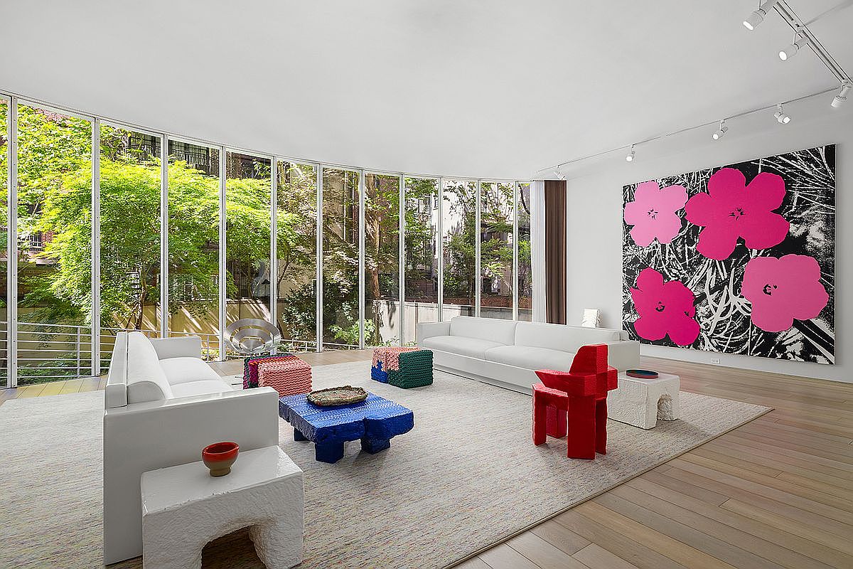 This expansive, modern living room features a curved wall of floor-to-ceiling windows that provide an immersive view of the lush, green trees outside. The space is furnished with two white sofas, a vibrant blue coffee table, and eclectic accent seating, all set atop a large, neutral-toned area rug. A prominent, large-scale floral art piece hangs on the wall, while track lighting mounted on the ceiling illuminates the bright, open-concept interior.