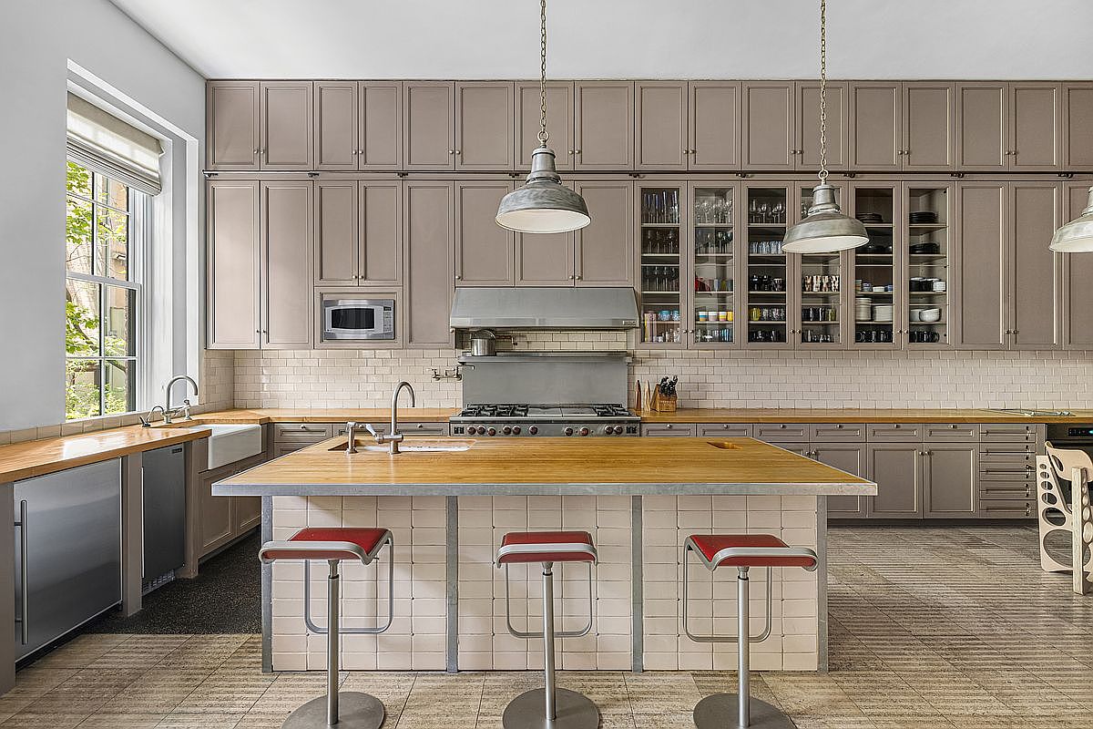 This spacious kitchen features a large central island with a wood countertop and three red-accented stools, set against a backdrop of extensive taupe cabinetry and a white subway tile backsplash. A professional-style range with a stainless steel hood serves as the focal point, complemented by a farmhouse-style sink and light wood countertops along the perimeter. The room is illuminated by industrial-style pendant lights and natural light streaming through a large window on the left, creating a functional and well-organized culinary space.